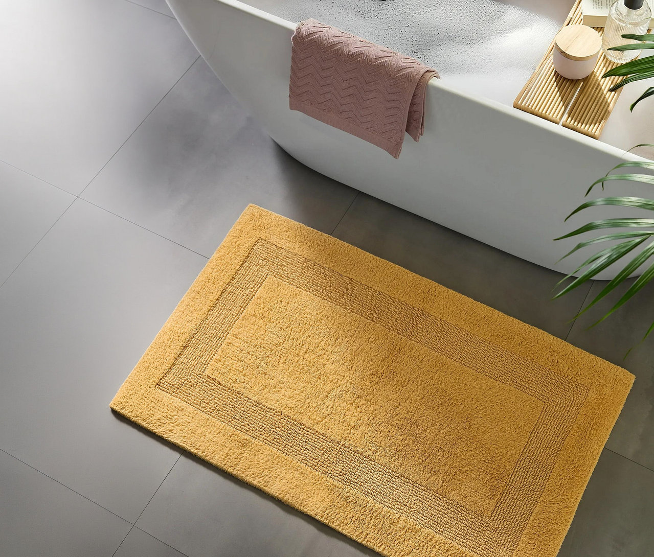 Rechteckige gelbe Badematte mit Bordürendesign auf grauem Fliesenboden, neben einer weißen Badewanne mit rosa Handtuch, einem Holzbadebrett und einer grünen Pflanze.