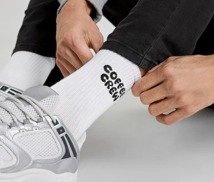 Mann zieht weiße Socken mit der Aufschrift „Coffee Crew“ und silberne Turnschuhe an.