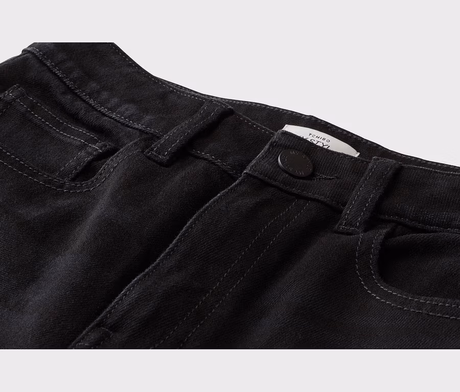 Nahaufnahme einer schwarzen Jeans mit vorderen Taschen, Knopf, Gürtelschlaufen und einem teilweise sichtbaren weißen Tchibo-Etikett.