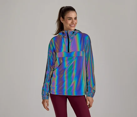 Lächelnde Frau trägt eine reflektierende, irisierende Anorak-Jacke mit Kapuze und halbem Reißverschluss, kombiniert mit dunkelroten Leggings.