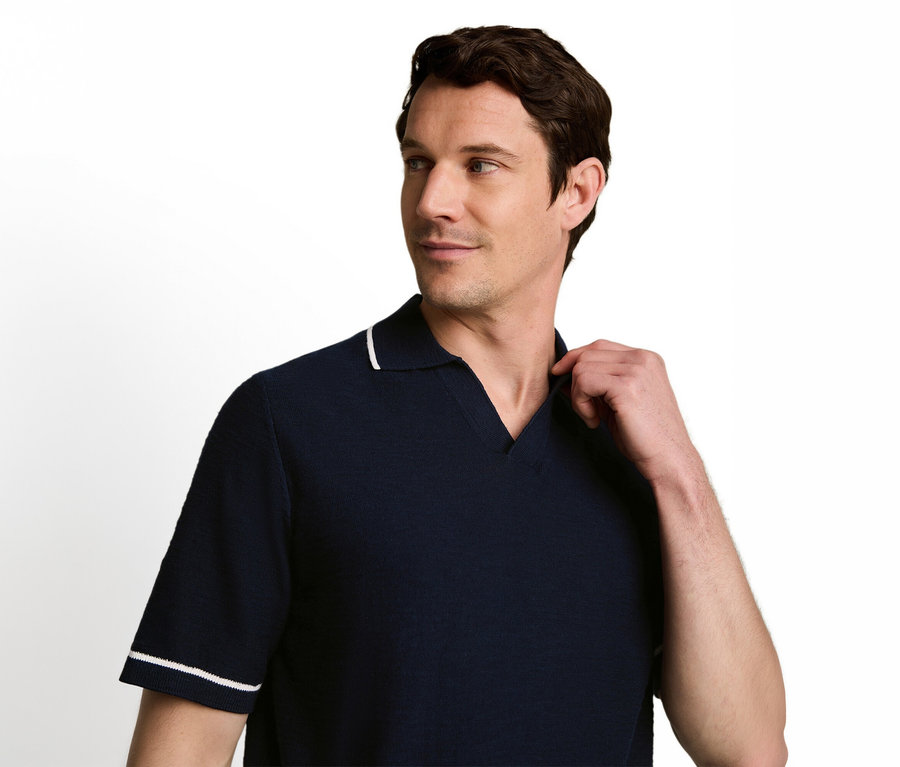 Ein Mann in einem dunkelblauen Poloshirt mit weiß gestreiftem Kragen und Ärmeln blickt nach rechts und berührt mit der linken Hand seinen Kragen.