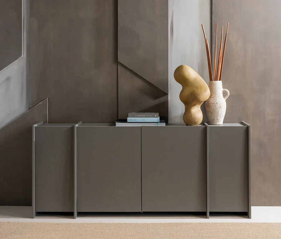 Ein modernes graues Sideboard mit Büchern, einer abstrakten Skulptur und einer Vase mit Stäben darauf, vor einer strukturierten grauen Wand und einem beigen Teppich.