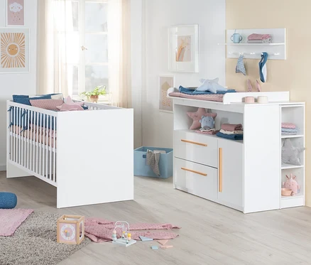 Ein Babyzimmer mit einem weißen Roba 2-teiligen Kinderzimmer-Set 2 »Lilo«, bestehend aus einem Gitterbett mit blauer und rosa Bettwäsche und einer passenden Wickelkommode mit Wickelauflage, Schubladen und offenen Fächern. Auf dem hellen Holzboden liegt ein grauer Teppich mit Holzspielzeug.