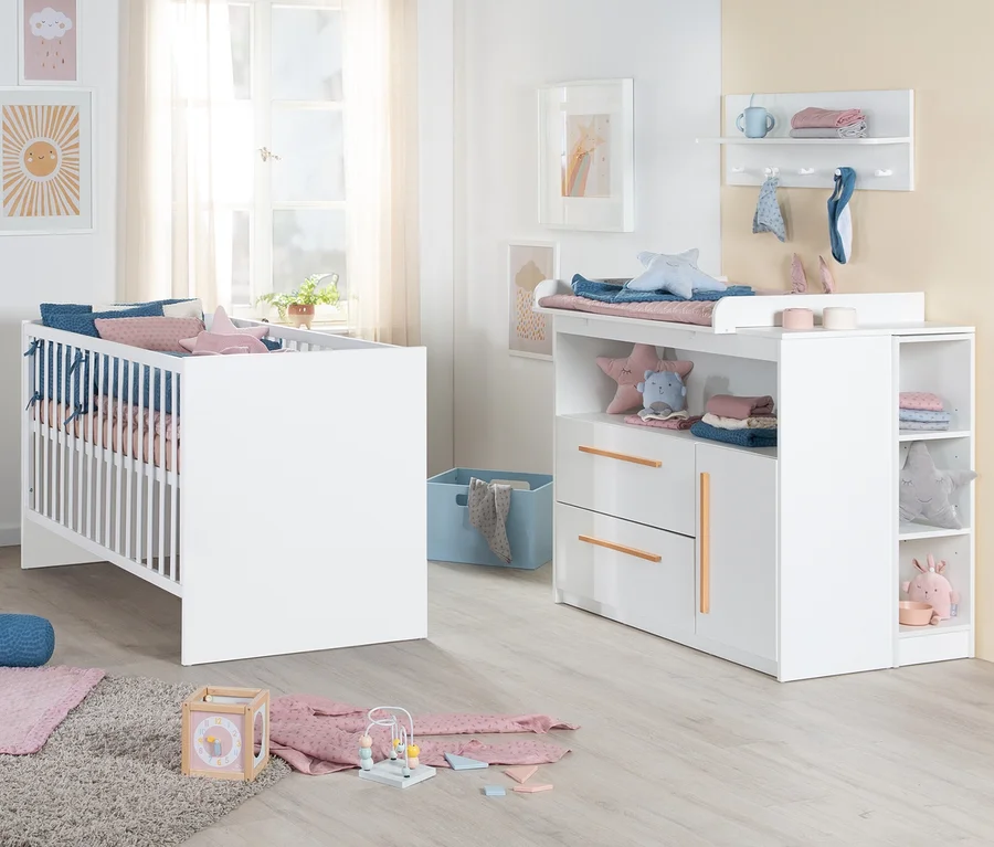 Ein Babyzimmer mit einem weißen Roba 2-teiligen Kinderzimmer-Set 2 »Lilo«, bestehend aus einem Gitterbett mit blauer und rosa Bettwäsche und einer passenden Wickelkommode mit Wickelauflage, Schubladen und offenen Fächern. Auf dem hellen Holzboden liegt ein grauer Teppich mit Holzspielzeug.