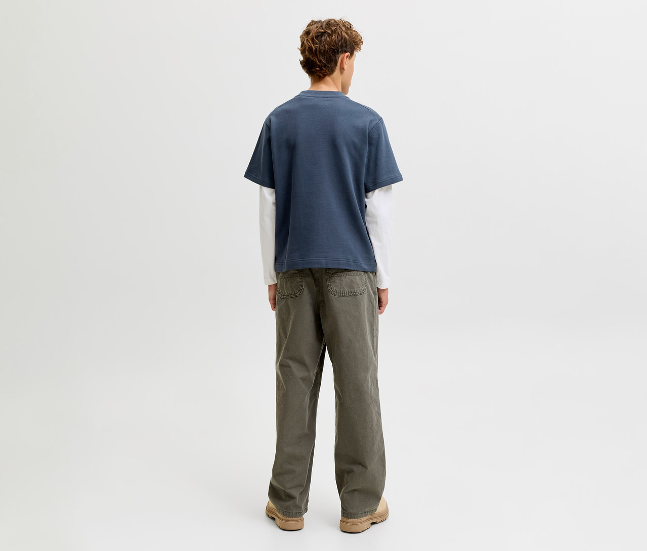 Eine Person steht mit dem Rücken zum Betrachter und trägt ein Jack&Jones double layered Shirt, bestehend aus einem blauen Kurzarmshirt über einem weißen Langarmshirt, dazu olivgrüne Hosen und hellbraune Schuhe.
