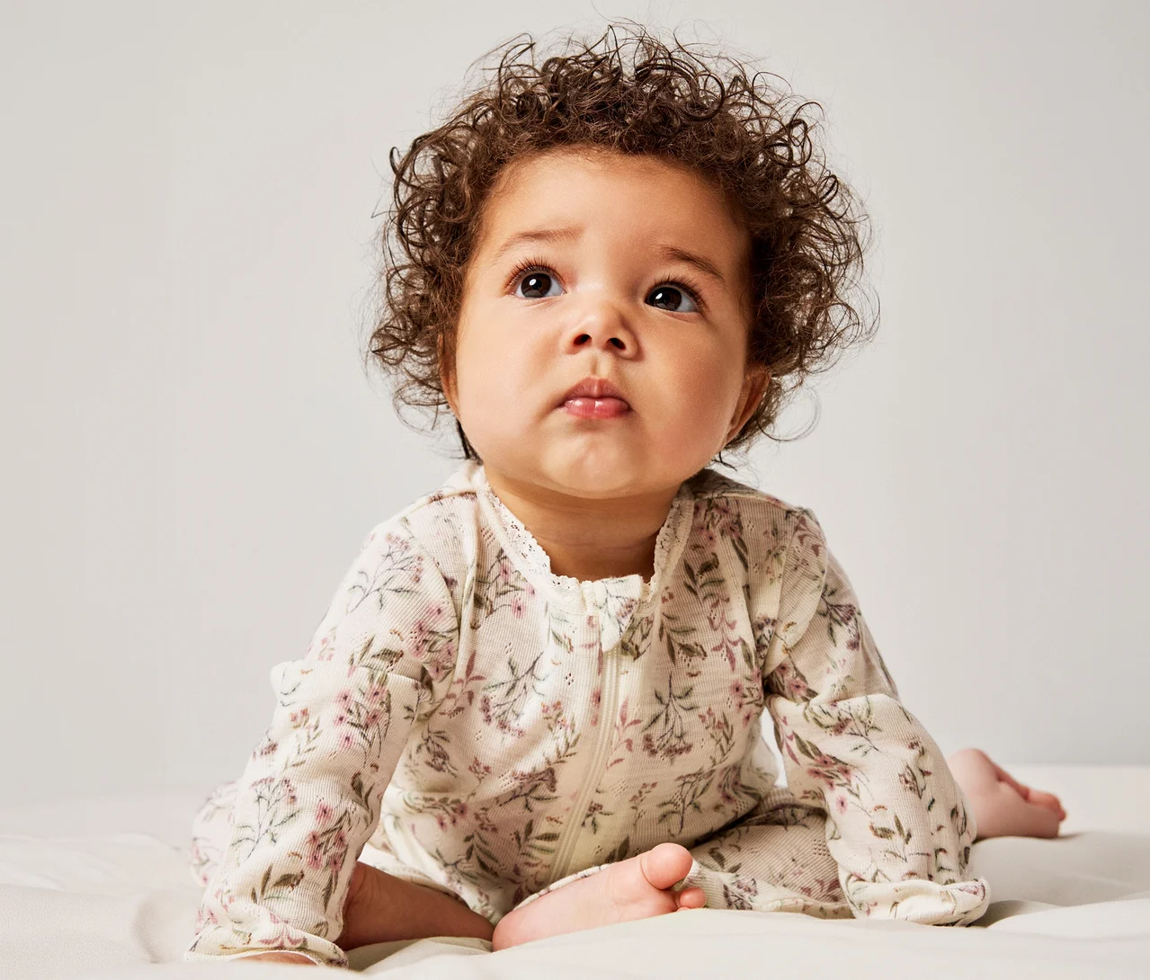 Baby mit lockigem braunem Haar liegt auf dem Bauch und blickt nach oben. Es trägt einen cremefarbenen, langärmeligen Strampler mit Blumenmuster, Reißverschluss und Spitzenbesatz.