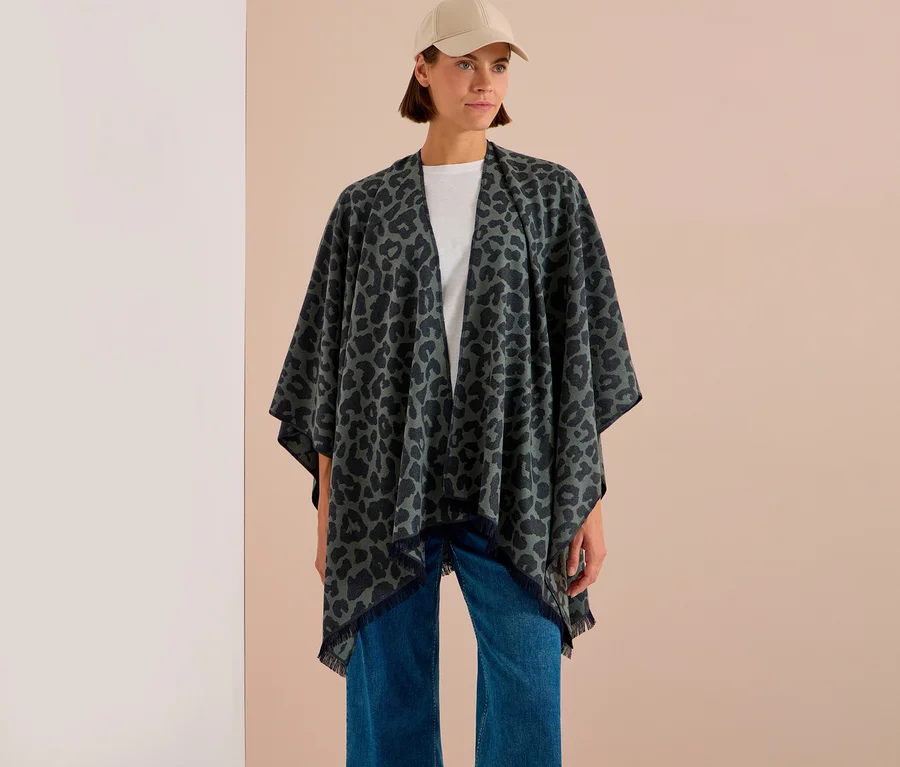 Frau trägt einen dunkelgrünen CECIL Poncho mit Leopardenmuster und Fransen, ein weißes T-Shirt, blaue Jeans und eine beigefarbene Kappe.