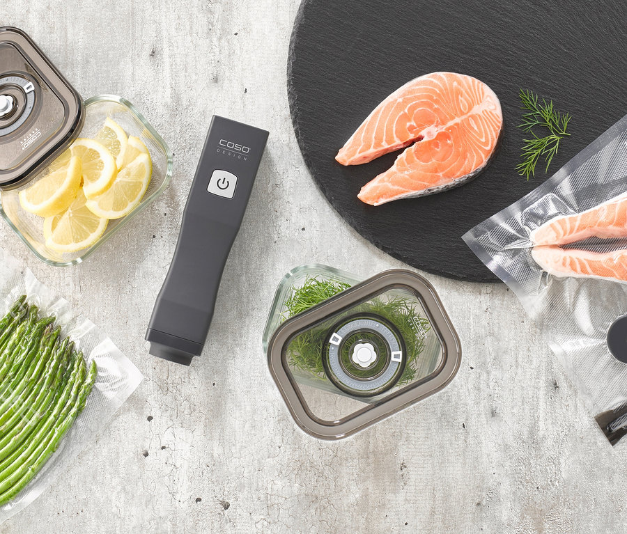 Ein dunkelgrauer CASO Vacu OneTouch Handvakuumierer liegt zwischen vakuumversiegelten Lebensmitteln: Lachsfilets, Zitronenscheiben in einem Behälter, Spargel in einem Beutel und Dill in einem weiteren Behälter.