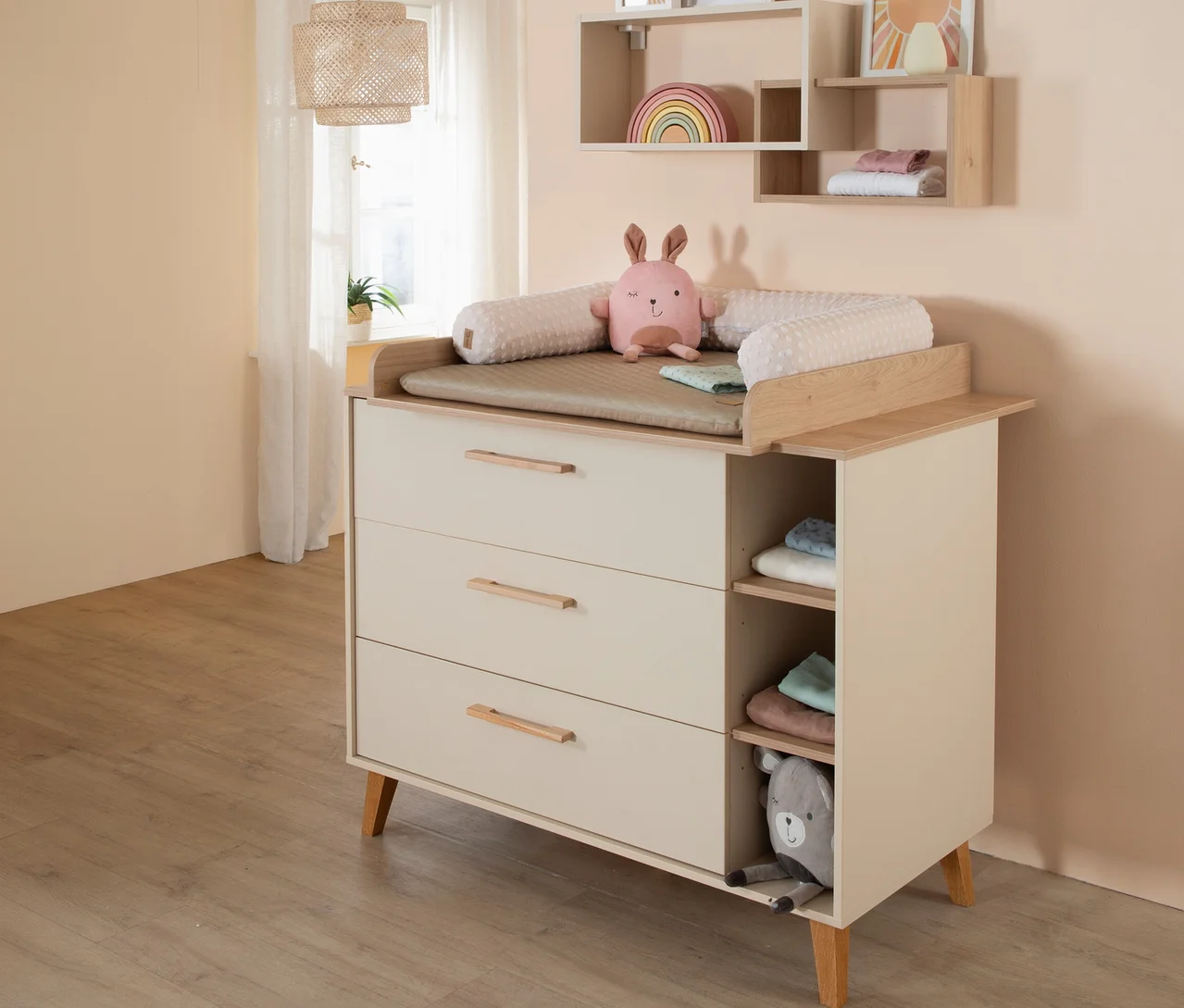 Cremefarbene Wickelkommode mit Holzgriffen und drei Schubladen. Oben liegen eine Wickelauflage, eine Nackenrolle und ein rosa Hasenkuscheltier. Rechts sind offene Fächer mit Babykleidung und einem grauen Bärenkuscheltier. Im Hintergrund sind Wandregale und ein Fenster zu sehen.
