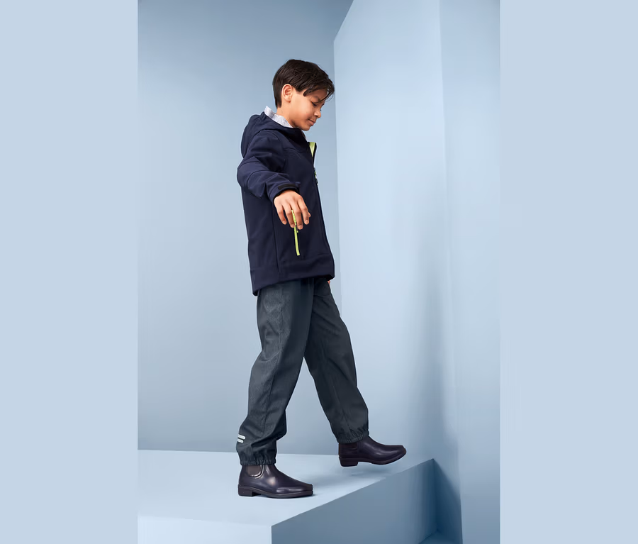 Ein Junge geht eine Stufe hoch, er trägt eine Softshellhose im Jeans-Look, eine Jacke und Stiefel.