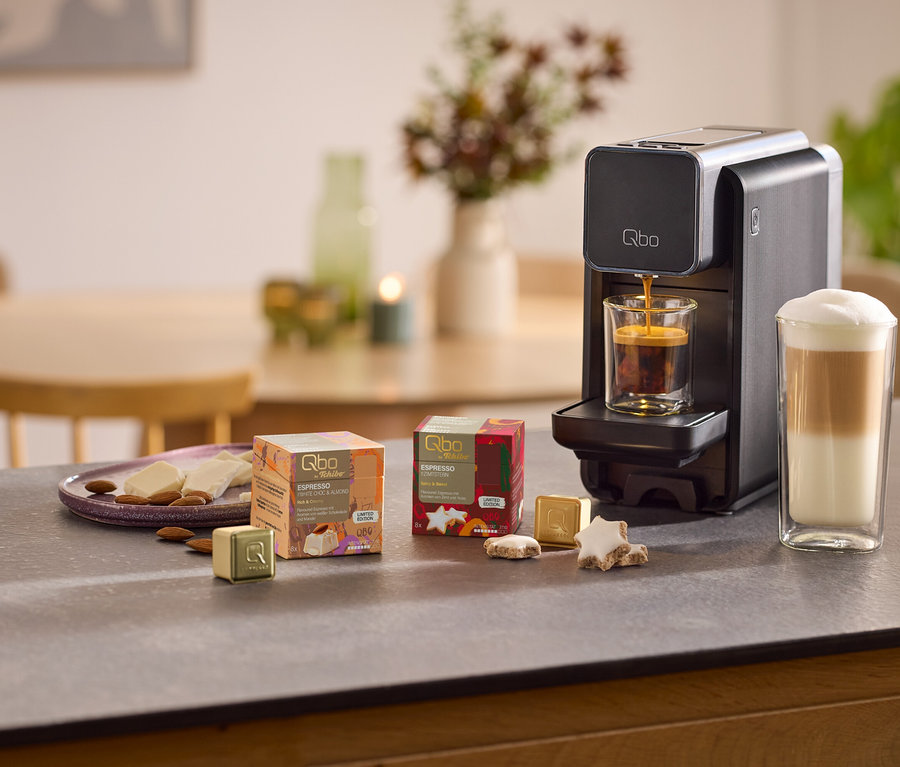 Eine schwarze Qbo ONE Kapselmaschine brüht Espresso in ein Glas, daneben ein Latte Macchiato. Auf der Arbeitsplatte liegen Qbo Espresso White Choc Mandel Packungen, goldene Kapseln, Sternkekse, weiße Schokolade und Mandeln.