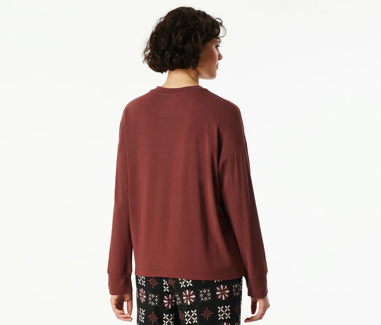 Frau von hinten, leicht gedreht, mit kurzen, dunklen Locken, trägt ein lockeres, rotbraunes Langarmshirt und eine schwarze gemusterte Hose mit weißen und rosa Blumenmustern.