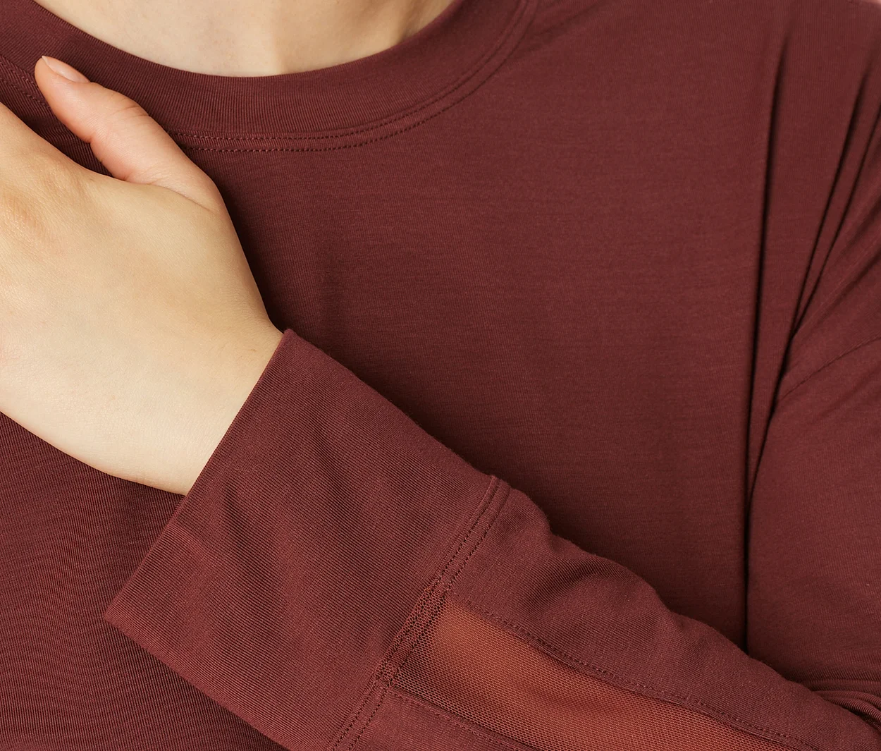 Nahaufnahme einer Person in einem bordeauxroten Langarmshirt mit Rundhalsausschnitt. Eine Hand ruht auf der Brust. Das Shirt hat dunklere Schulterpartien und einen Netzeinsatz am Unterarm.