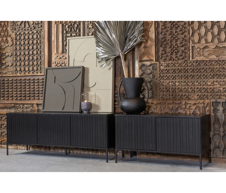 Zwei schwarze Holz-Sideboards mit Lamellentüren stehen vor einer Wand aus geschnitzten Holzpaneelen. Auf dem linken Sideboard sind zwei abstrakte Kunstwerke und eine Lampe, auf dem rechten eine schwarze Vase mit einem silbernen Palmenblatt.