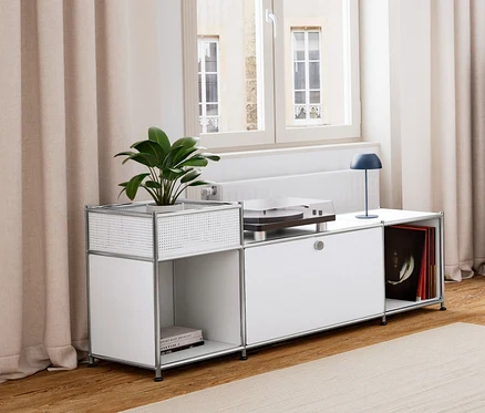 Ein Wohnzimmer mit Holzboden und hellem Teppich. Links steht ein weißes Lowboard Metall »CN3« mit Klappenfach, darauf eine dunkelblaue LED-Akku-Tischleuchte. Daneben eine dunkelblaue Kommode Metall »CN3« mit 3 Klappenfächern. In der Mitte ein Beistelltisch mit Tablett und Sitzfunktion und ein Sessel. Im Hintergrund ein hellgraues Sideboard Metall »CN3« mit 3 Klappenfächern.
