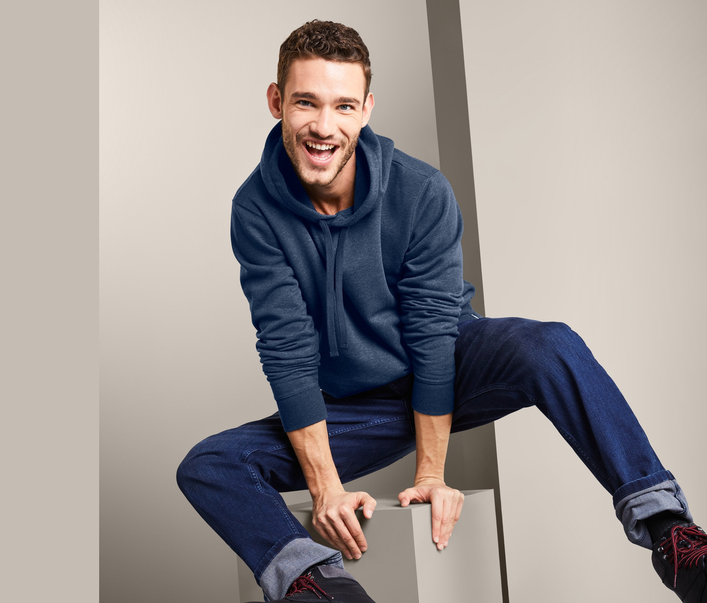 Lächelnder Mann sitzt auf einem grauen Würfel, trägt einen blauen Hoodie und Jeans.