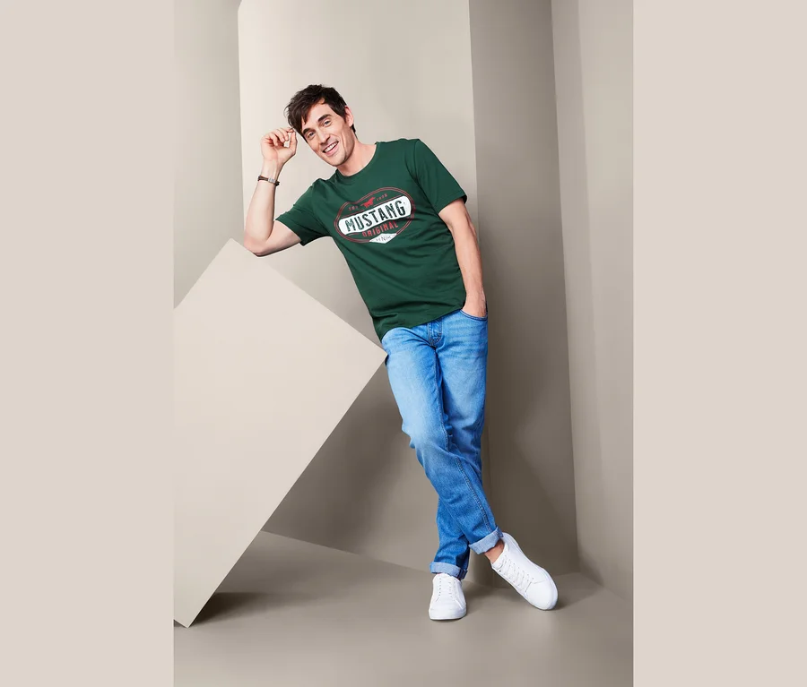 Mann posiert an eine Wand gelehnt in einem grünen T-Shirt »Mustang«, blauen Jeans und weißen Turnschuhen.