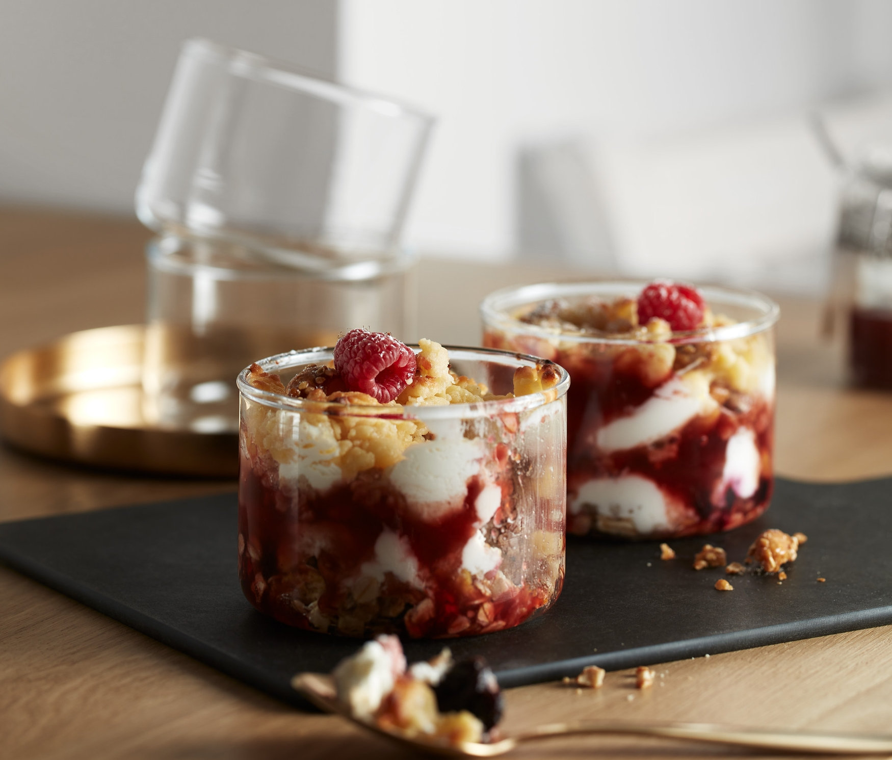 Auf einem Tisch stehen 4 Portions-und-Ofengläser, gefüllt mit einem Fruchtdessert. Zwei Gläser stehen auf einem schwarzen Teller, zwei weitere sind auf einem goldenen Tablett gestapelt.