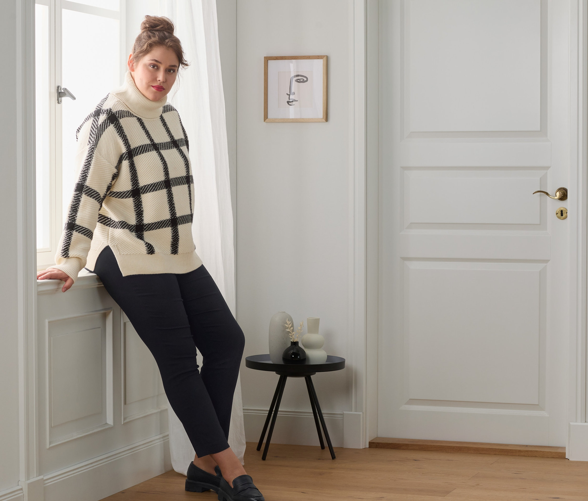 Eine Frau lehnt am Fenster und trägt einen Strickpullover mit Rollkragen, eine Stretchhose und Leder-Loafer.