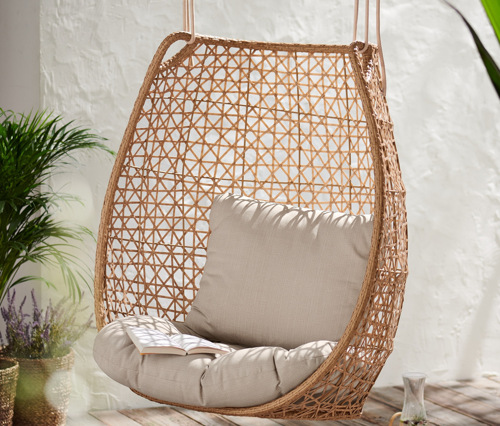 Ein Hängesessel »Boho« mit beigem Kissen und Buch auf einem Holzboden.