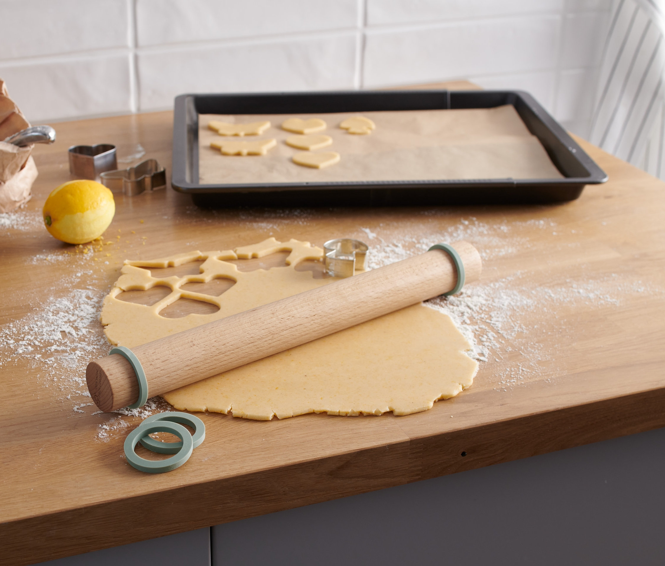 Auf einer Holzfläche liegt eine Holz-Teigrolle neben ausgerolltem Teig mit ausgeschnittenen Formen. Auf einem Backblech liegen ausgeschnittene Teigformen.