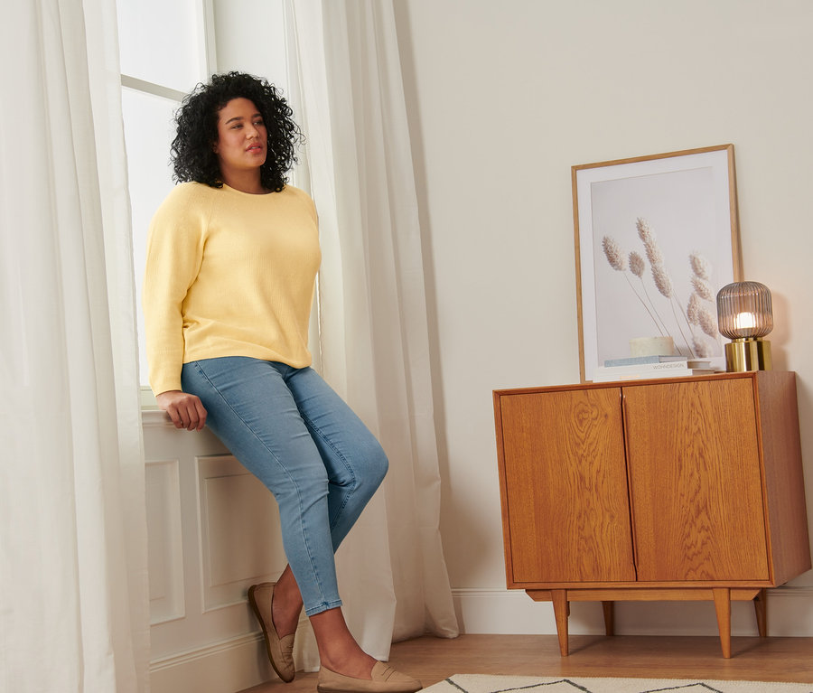 Frau sitzt auf Fensterbank in gelbem Strickpullover, Treggings und Loafer aus Veloursleder.