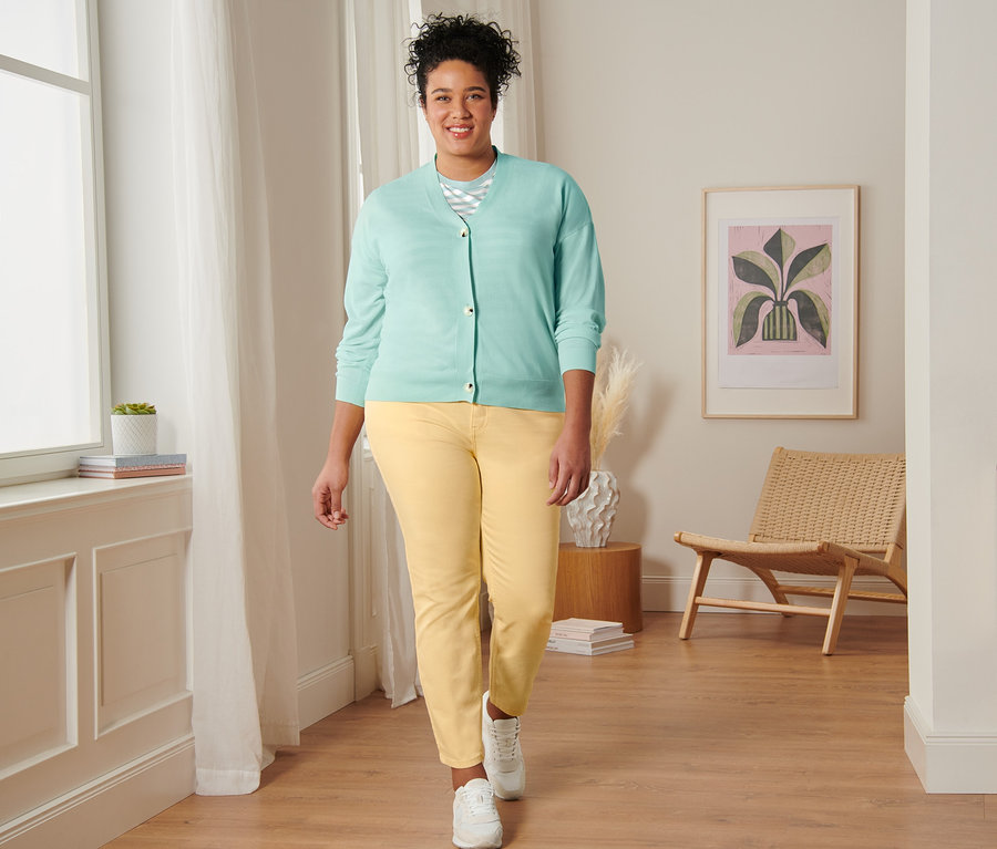 Eine Frau geht und trägt ein mintfarbenes, gestreiftes T-Shirt, einen Cardigan mit V-Ausschnitt, eine gelbe Slimfit-Jeans und Veloursleder-Sneaker.