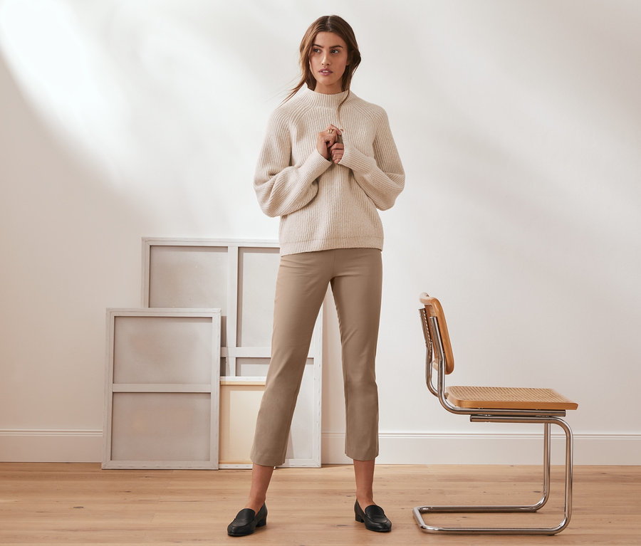 Frau steht und trägt eine 7/8-Stretch-Hose in Camel, einen cremefarbenen Pullover und schwarze Slipper.