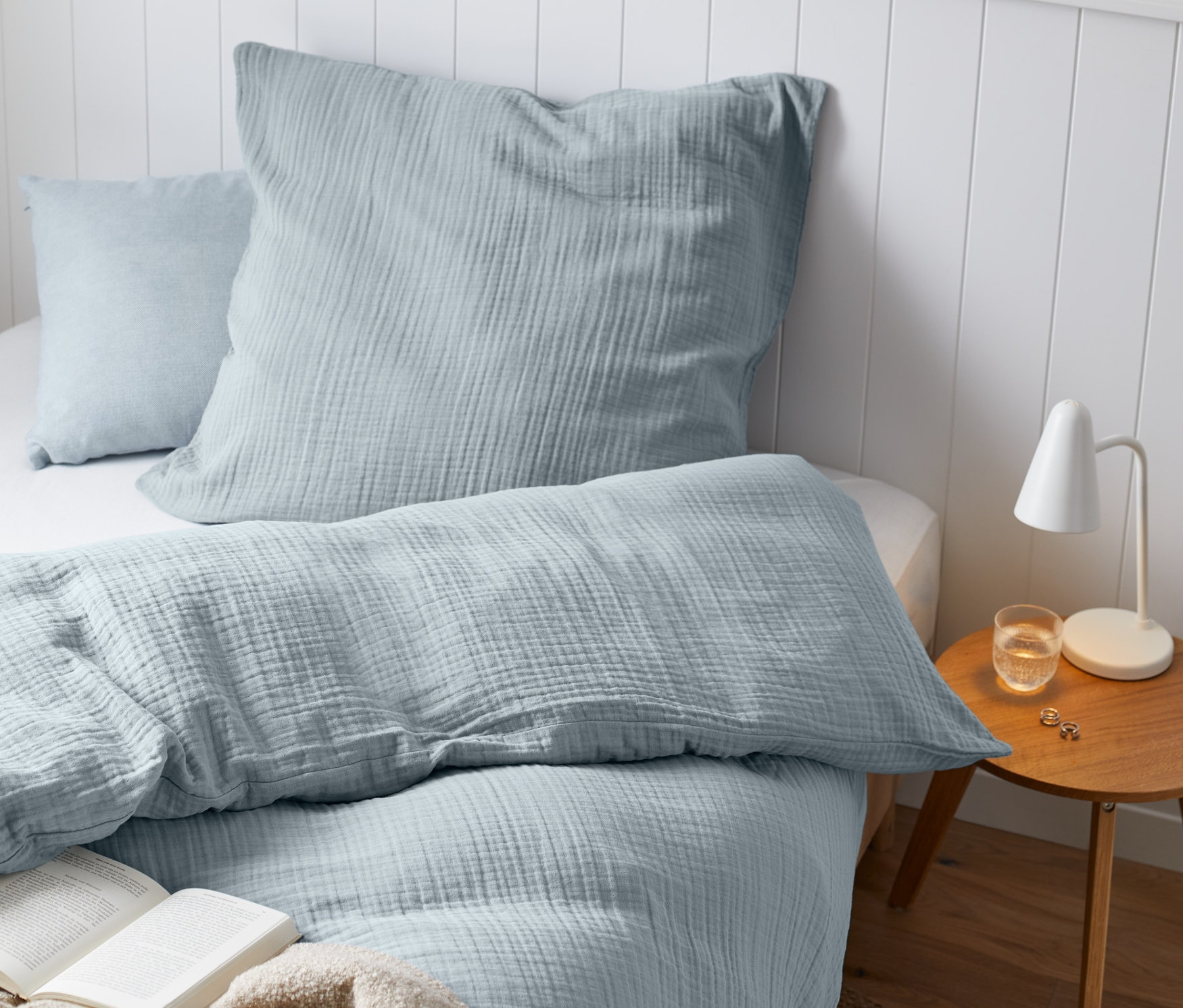 Hellblaue Musselin-Bettwäsche auf einem Bett mit einem offenen Buch und einer beigen Decke.