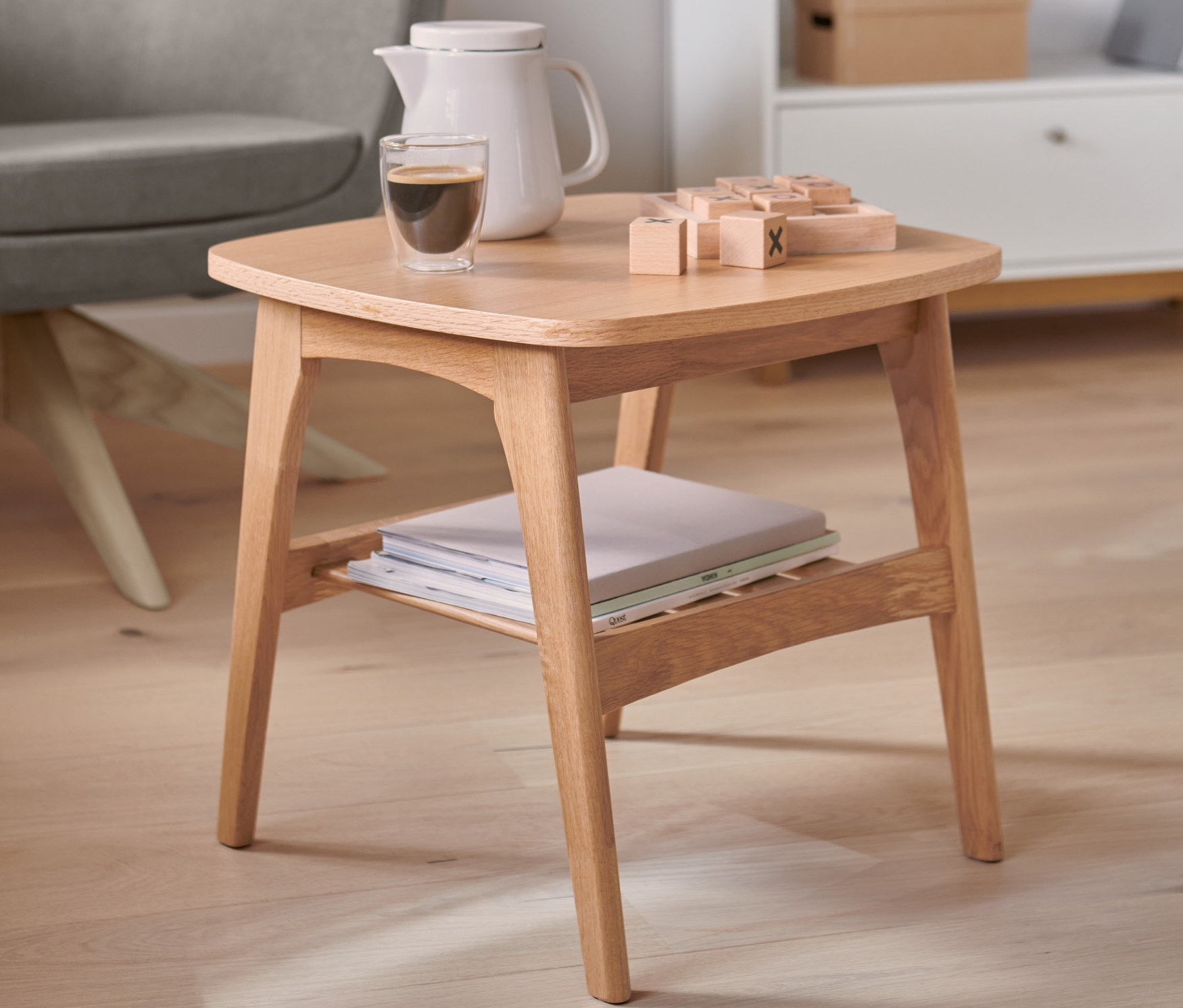 Auf einem Holztisch stehen eine Kaffeekanne, ein Glas Kaffee und Holzklötze. Im Hintergrund sind ein grauer Sessel und ein weißes Bücherregal zu sehen.