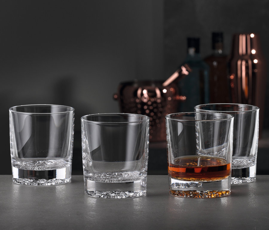 Vier Gläser stehen auf einem Tisch, eines davon ist mit einer braunen Flüssigkeit gefüllt. Im Hintergrund sind Flaschen und Barzubehör zu sehen. Dargestellt ist ein Spiegelau Whiskybecher »Lounge 2.0«, 8er-Set.