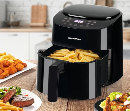 Auf einem Holztisch steht eine schwarze digitale Heißluftfritteuse mit Pommes frites darin sowie Teller mit Chicken Nuggets, Pommes frites, Steak und Salat.