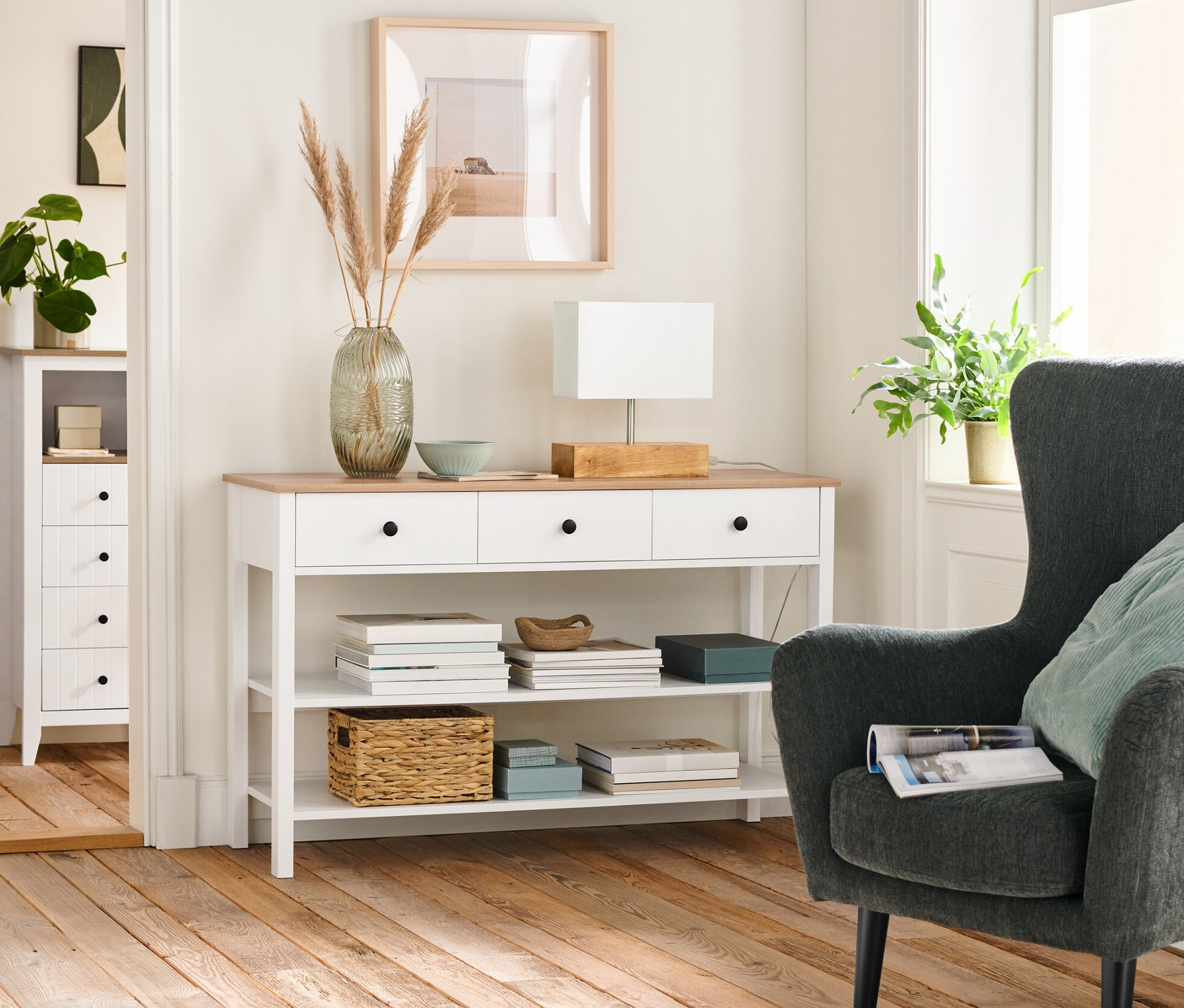 Wohnzimmer Interieur mit einem anthrazitfarbenen, gepolsterten Sessel, einer Anrichte mit Schubladen und einer Tischleuchte mit Holzfuß.