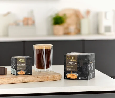 Auf einem Tisch stehen eine Tasse Kaffee und Packungen Qbo Caffè Grande ESPÍRITO SÃO PAULO mit 8 und 27 Kapseln.
