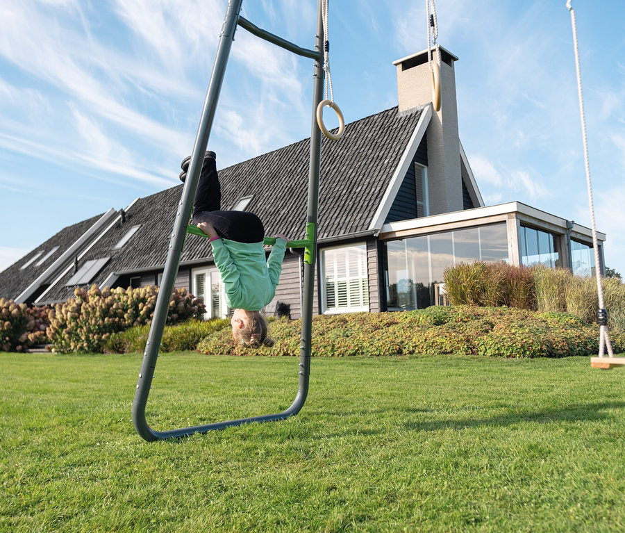 Mädchen hängt kopfüber an grauem EXIT Klettergerüst MB210 auf einer Rasenfläche vor einem Haus.
