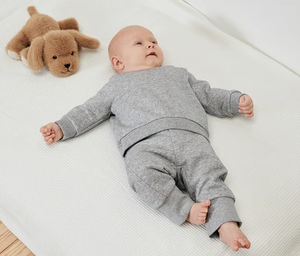Baby liegt auf einer weißen, strukturierten Decke neben einem Stoffhund und trägt eine graue 2 Baby-Sweathosen mit angerauter Innenseite.