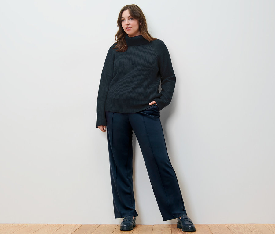 Eine Frau posiert auf einem Holzboden in einem dunkelblauen Grobstrickpullover mit Rollkragen und einer Jerseyhose mit Biese.