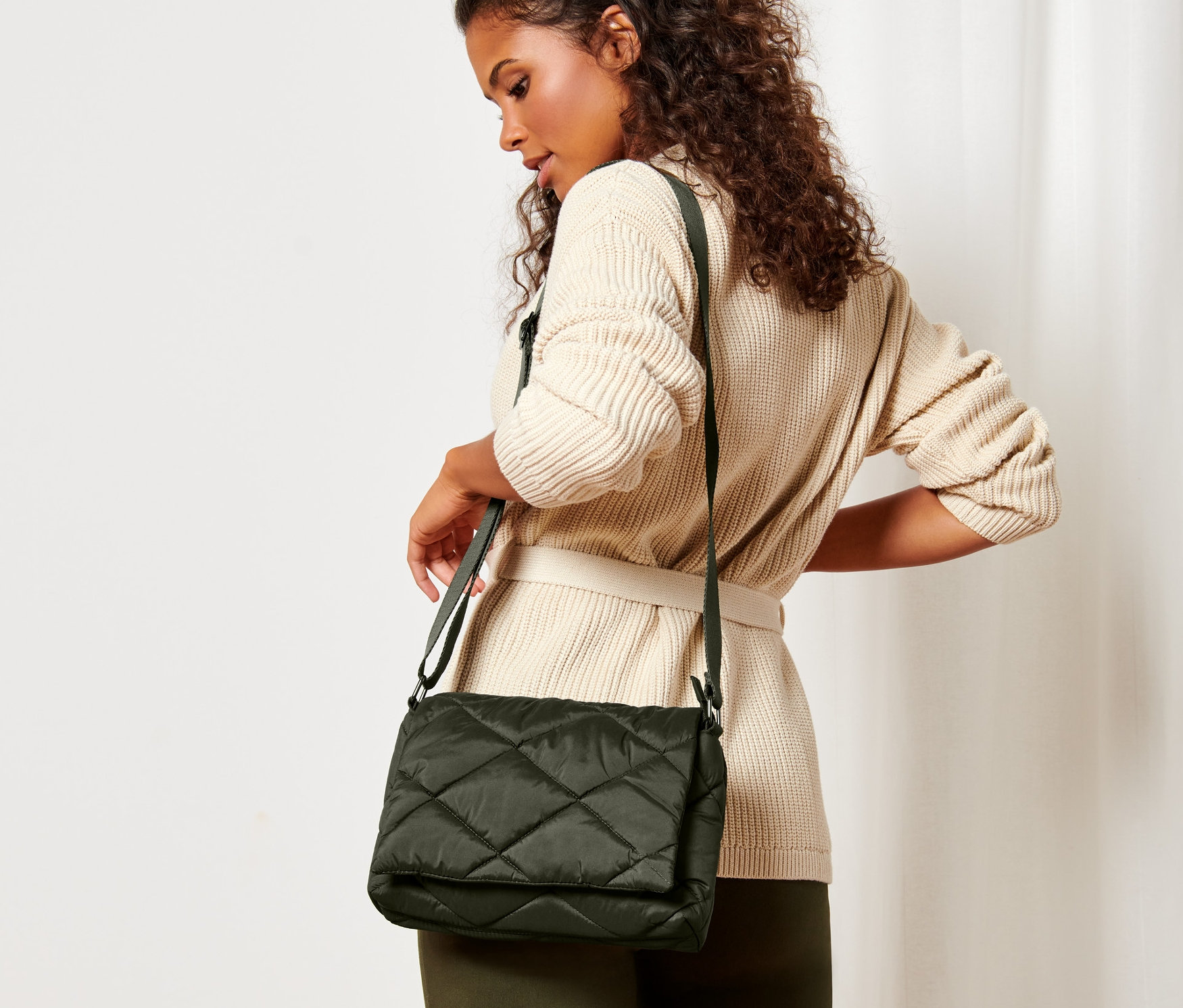 Frau mit braunen Locken und beigem Gürtel-Cardigan posiert mit gesteppter Umhängetasche.