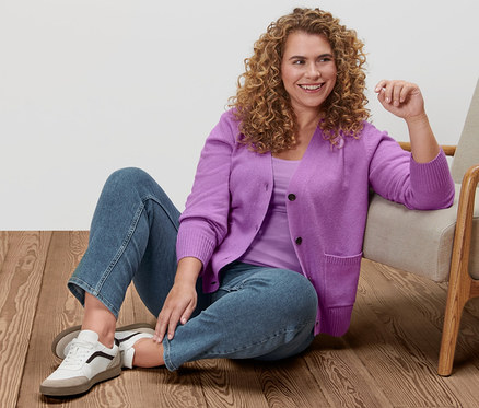 Frau sitzt auf dem Boden neben einem Sessel und trägt eine Strickjacke, ein Hemdchen, Straight-Denim und Leder-Sneaker.