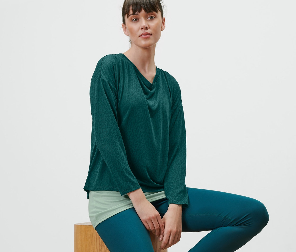 Frau sitzt auf einem Holzhocker und trägt ein grünes 2-in-1-Sportshirt.