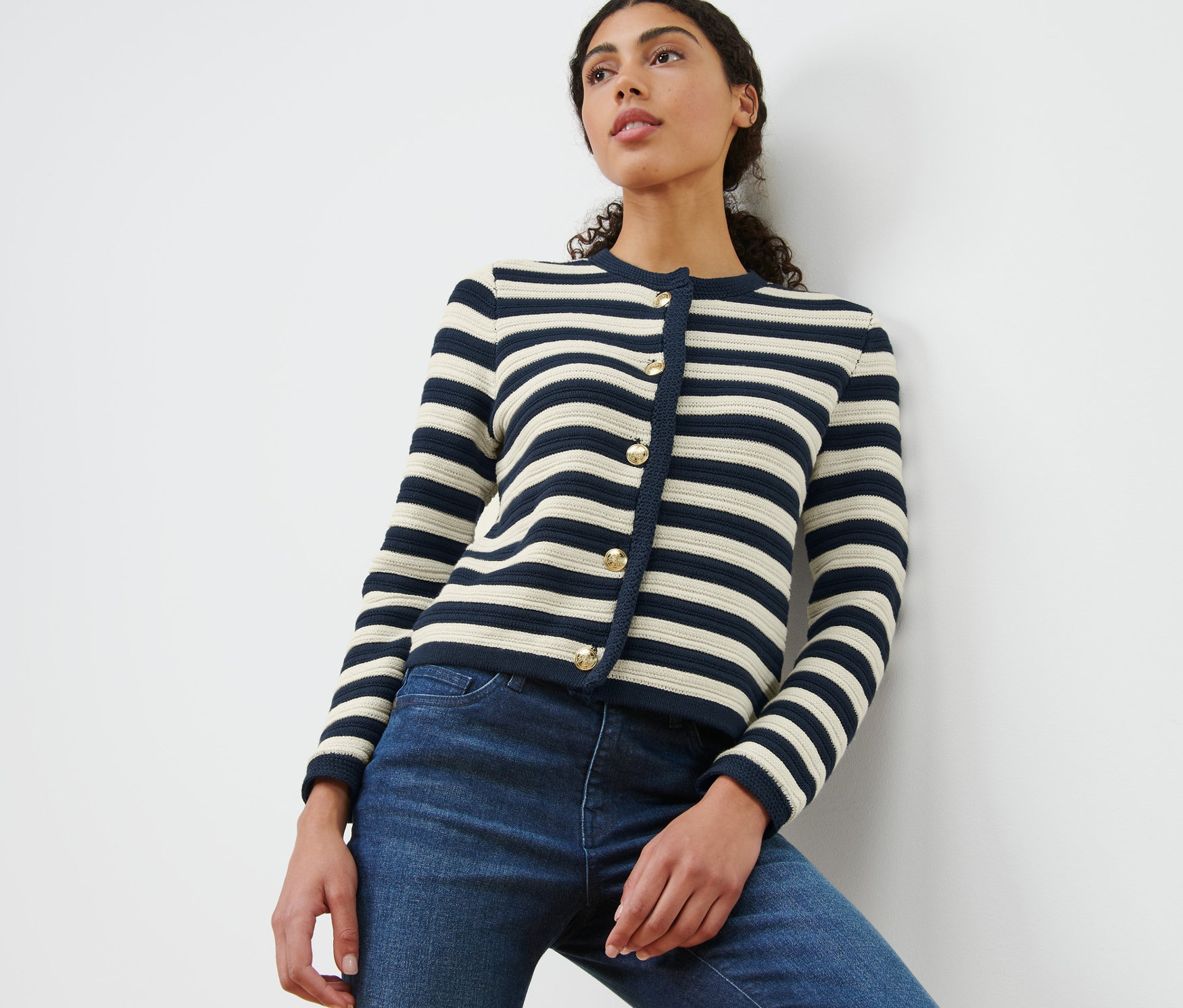 Eine Frau, die sich an eine weiße Wand lehnt, trägt eine Cardigan aus Strukturstrick mit marineblauen und cremefarbenen Streifen und Jeans.