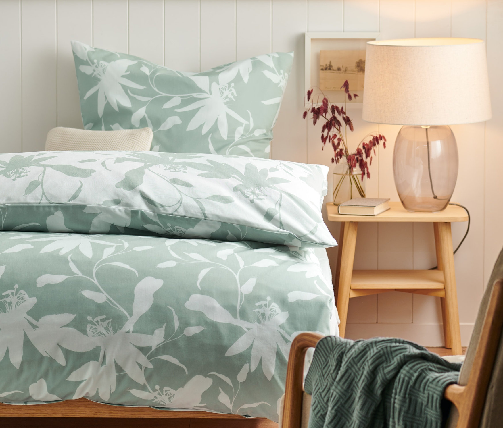 Bett mit Premium-Baumwoll-Bettwäsche mit Blumenmuster, Nachttisch mit Lampe und Sessel mit Decke.