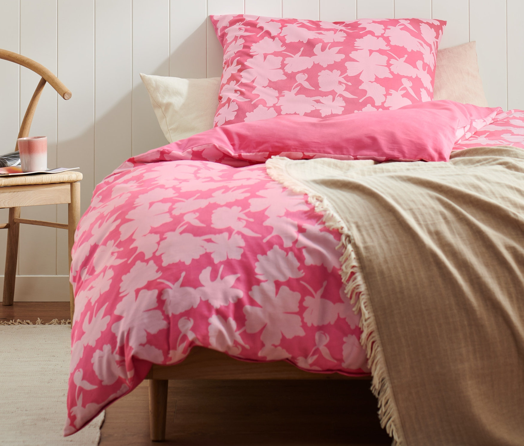 Bett mit rosa Premium-Baumwoll-Bettwäsche mit Blumenmuster in Normal- und Übergröße. Auf dem Nachttisch steht eine Kerze, auf dem Stuhl liegt eine Zeitschrift.
