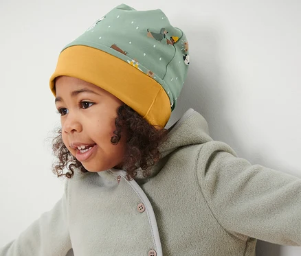 Mädchen mit lockigem Haar trägt eine grün-gelbe Kinder-Wendemütze und eine grüne Jacke.