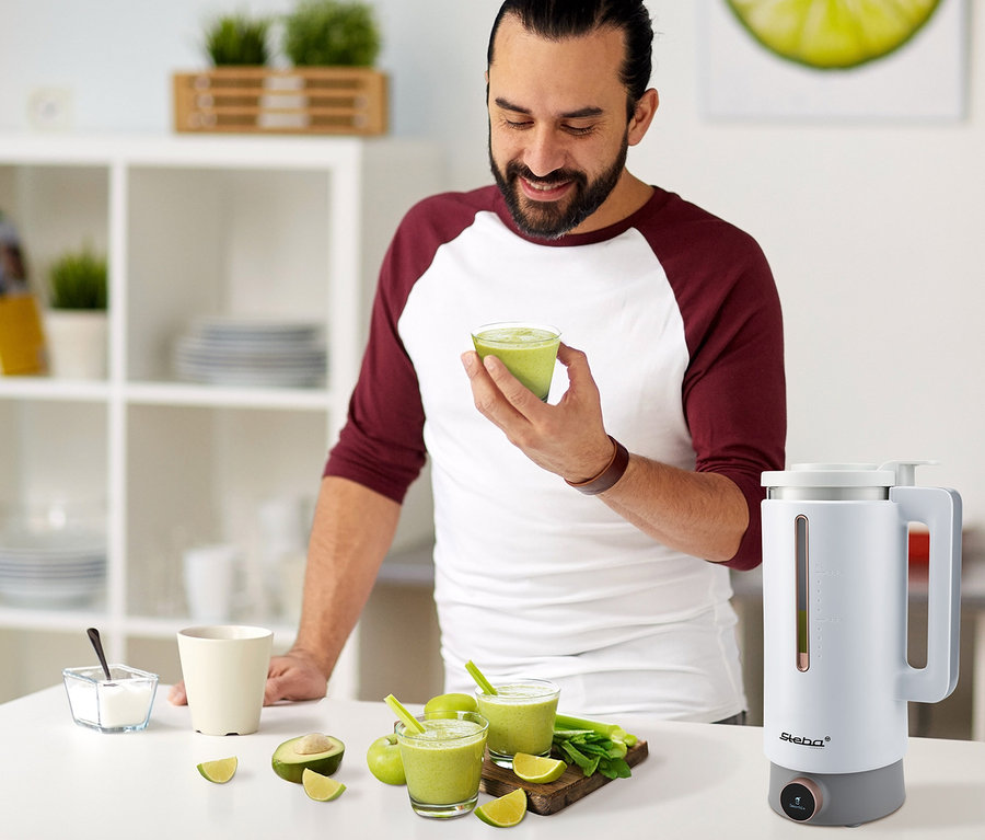 Mann hält ein Glas grünen Smoothie. Auf dem Tisch stehen Steba Vegan-Drink-Maker "VDM 2 HOT & COLD", Gläser, Früchte und Smoothies.