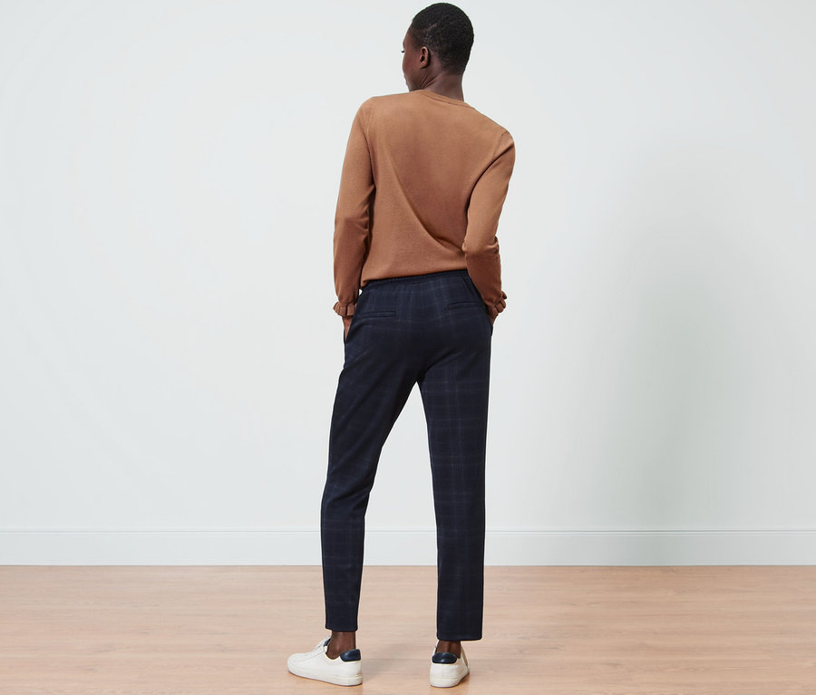 Person von hinten stehend, trägt eine karierte Joggpants, einen braunen Pullover und weiße Turnschuhe.