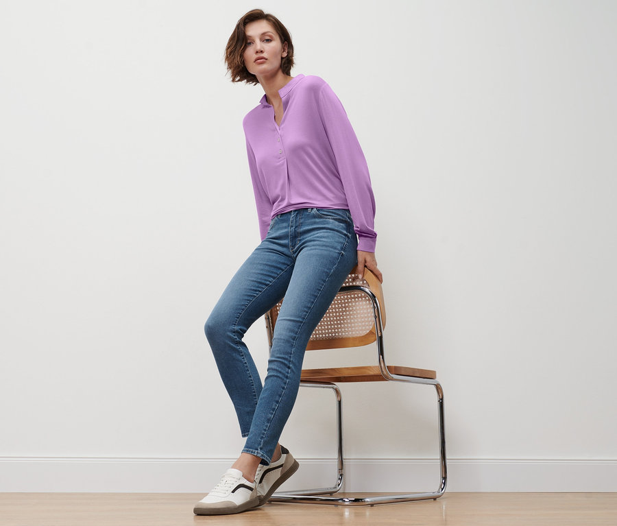 Eine Frau sitzt auf einem Stuhl und trägt Slim-Denim »Fit Emma« in Midblue, ein fliederfarbenes Blusenshirt mit Knopfleiste und Leder-Sneaker.
