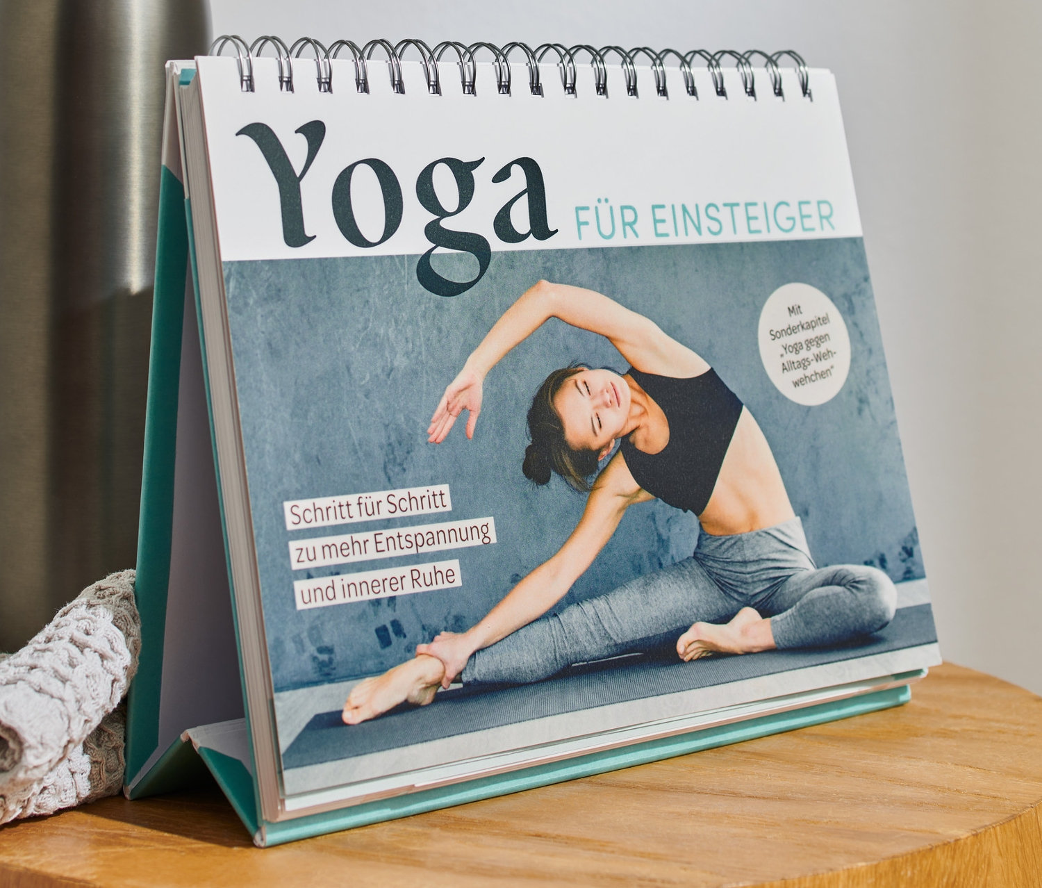 Auf einem Holztisch liegen ein »Yoga für Einsteiger« Buch, eine Edelstahlflasche und ein graues Stricktuch.