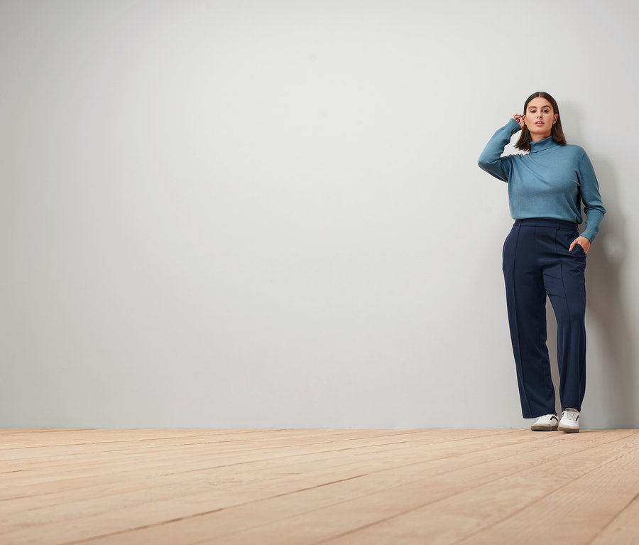 Frau posiert in rauchblauem Feinstrick-Rollkragenpullover, dunkelblauer Jerseyhose mit Biese und Leder-Sneakern.