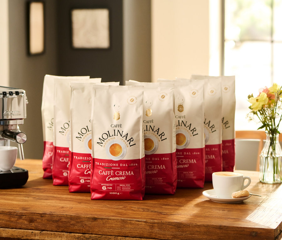 Auf einem Holztisch stehen eine Espressomaschine neben einer Tasse mit Kaffee und einem Keks. Dahinter befinden sich acht Packungen Caffè Molinari Caffè Crema Cremoso - 8x 1 kg Ganze Bohne und eine Vase mit Blumen.
